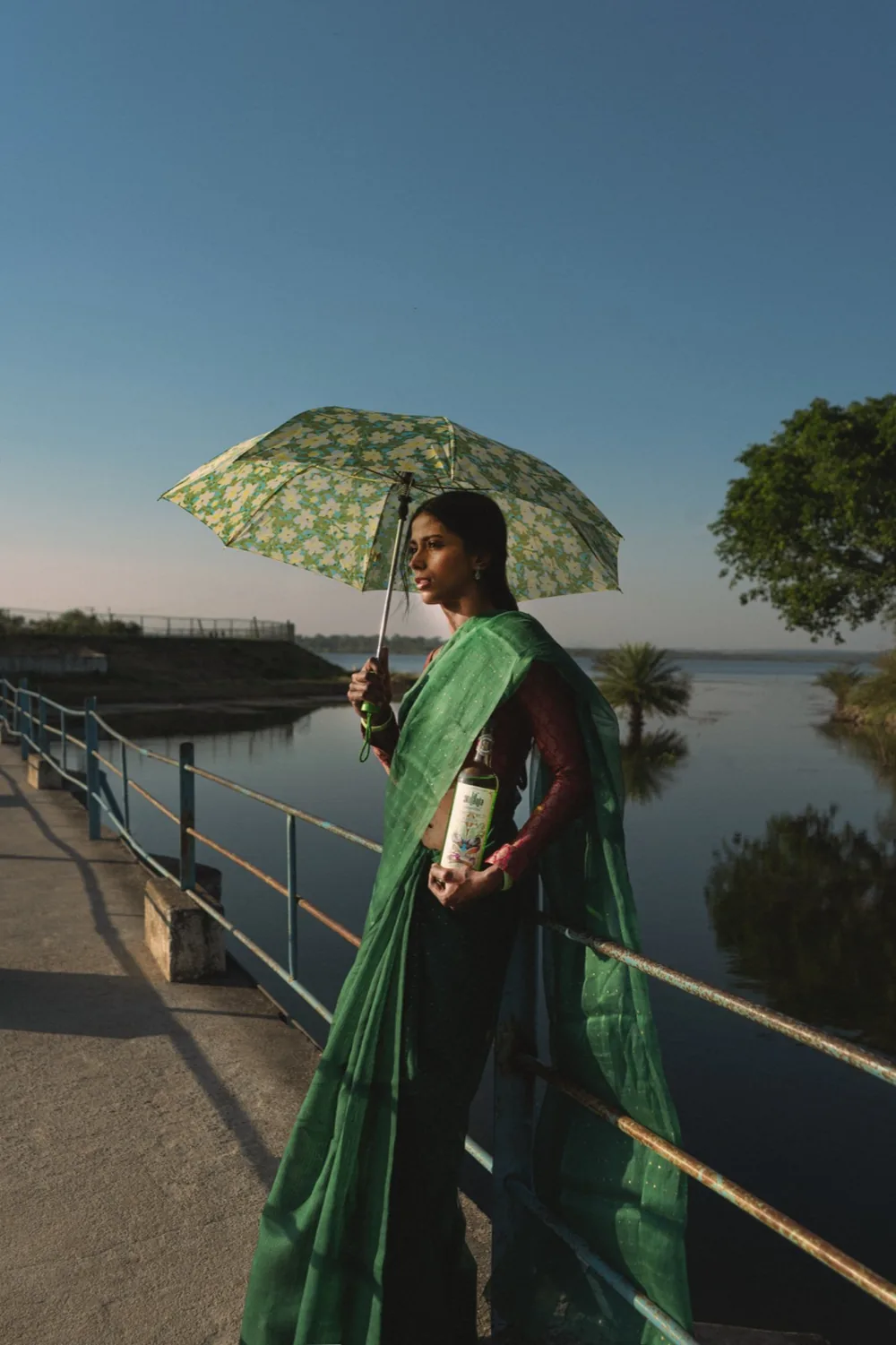 A woman in a green sari holding a bottle of La'Ananta Absinthe under an umbrella by a lake at dusk