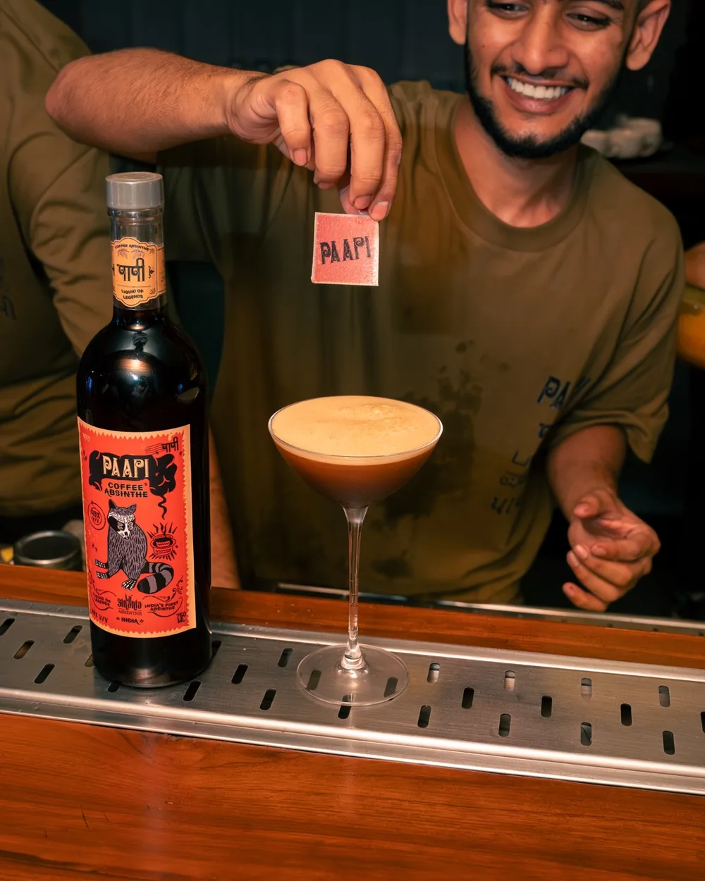 A bartender holding a Paapi tag over a frothy cocktail beside a bottle of Paapi Coffee Absinthe